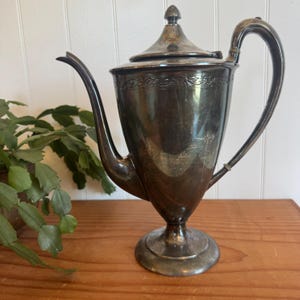 May include: A vintage silver-plated coffee pot with a long, curved spout and a curved handle. The pot has a lid with a decorative finial and a floral design around the top. It sits on a pedestal base.