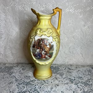 May include: Yellow ceramic pitcher with a handle and a spout. The pitcher features a painted scene of figures in classical attire within an ornate frame. The pitcher has a gold-colored handle and trim.