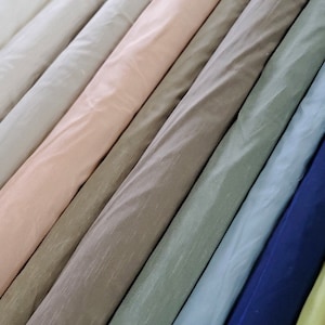 Puede incluir: Una colección de rollos de tela en varios colores, incluyendo blanco, rosa, beige, verde, azul y azul marino. La tela tiene un brillo sutil y una superficie ligeramente texturizada.