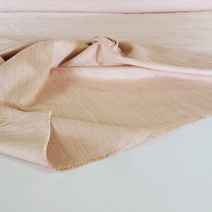 May include: A close-up of a pale peach-coloured fabric with a subtle, textured, woven pattern. The fabric is folded and draped, showing its soft, flowing texture.