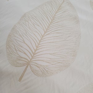 Op de afbeelding: Een witte stof met een reliëf beige bladontwerp.