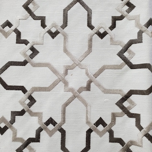 May include: Close-up of a white fabric with an embroidered geometric pattern. The design features interlocking shapes in shades of brown and tan, creating a symmetrical, repeating motif. The texture of the fabric is visible, adding depth to the design.
