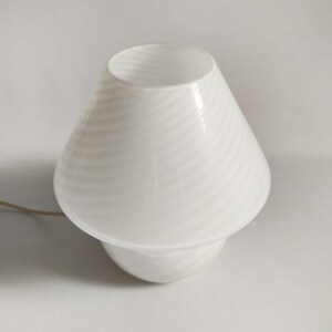 Pu&ograve; includere: Lampada da tavolo in vetro bianco con paralume conico e motivo testurizzato a spirale. La lampada ha un cavo bianco.