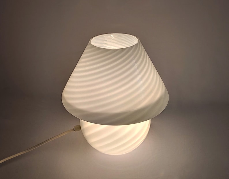 Pu&ograve; includere: Una lampada da tavolo bianca con un paralume e una base in vetro a spirale con texture. La lampada &egrave; accesa e illuminata.