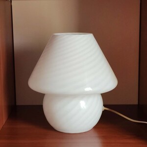 Pu&ograve; includere: Lampada da tavolo bianca a forma di fungo con un cavo bianco. La lampada ha un paralume e una base bianchi con un motivo a spirale.