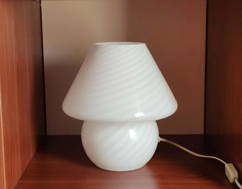 Pu&ograve; includere: Lampada da tavolo bianca a forma di fungo con un cavo bianco. La lampada ha un paralume e una base bianchi con un motivo a spirale.