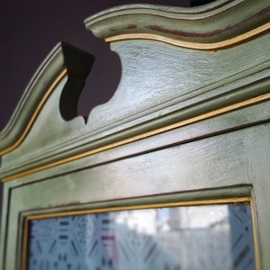 May include: A green wooden cabinet with gold trim and a glass door. The cabinet has a decorative top with a curved design.