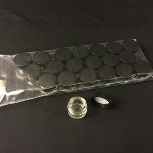 Könnte beinhalten: Ein klares Glasgefäß mit einem schwarzen Deckel. Das Gefäß ist leer und der Deckel ist geschlossen. Hinter dem Gefäß befinden sich 24 schwarze Deckel in einem Plastikbeutel.