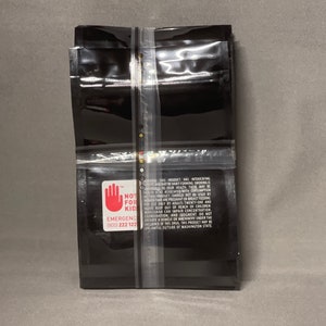 May include: A stack of black plastic bags with a warning label that reads "NOT FOR KID" and includes an emergency contact number. The label also includes a red hand with a stop sign.