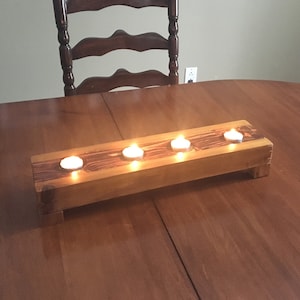 Puede incluir: Un portavelas de madera con cuatro velas de té encendidas. El soporte rectangular es de madera de color marrón claro y se encuentra sobre una mesa de madera marrón oscuro. El fondo incluye una silla de madera y fotos enmarcadas.