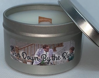 Puede incluir: Una lata de vela plateada con una vela blanca en su interior. La etiqueta de la lata muestra una escena de la película "Van Down By the River" con el texto "Van Down By the River".