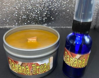 May include: A silver candle tin with a brown wax candle and a wooden wick. The tin has a label that says "Banana Bread". Next to the candle is a small blue glass bottle with a spray nozzle and a label that says "Banana".