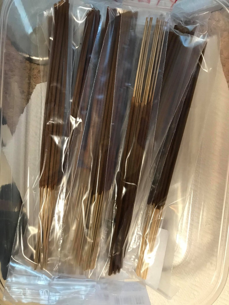 May include: Multiple bundles of brown incense sticks are packaged in clear plastic bags. The bags are stacked in a clear plastic container.