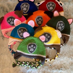 May include: A set of eight crocheted hats featuring the Paw Patrol characters. The hats are in various colours, including blue, red, pink, orange, teal, yellow, green, and brown. Each hat has a paw print emblem and two floppy ears.