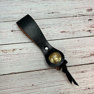 May include: Black leather key chain with a brass compass charm. The compass features a circular design with a white centre and black markings. The key chain has a tassel.
