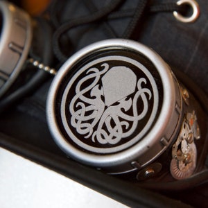 May include: A silver-colored metal steampunk-style goggles with a black and silver design of a stylized octopus with tentacles. The goggles have a chain attached to the side.