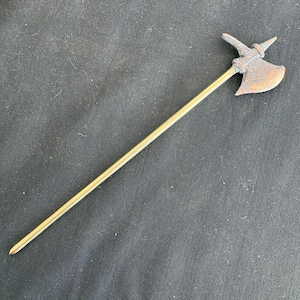 May include: A miniature bronze-colored halberd with a long, slender, gold-colored shaft. The axe head features a curved blade and a pointed spike. The halberd is a historical weapon, likely a decorative item.