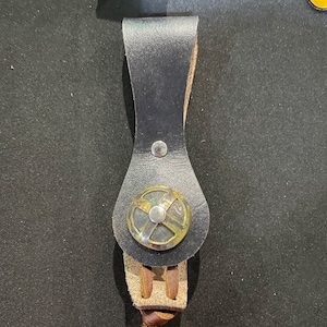 May include: A black leather strap with a metal buckle. The buckle has a circular design with a cross pattern.