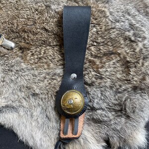 May include: A black leather strap with a brass button and a silver rivet. The button features a bird design. The strap is attached to a piece of light brown leather. The background is a furry, grey and brown surface.