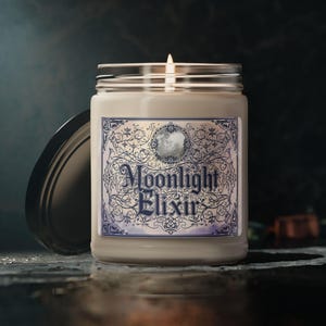 May include: A lit candle in a glass jar with a silver lid. The label reads "Moonlight Elixir" in elegant script, surrounded by a blue and white floral design. The candle sits on a dark surface.
