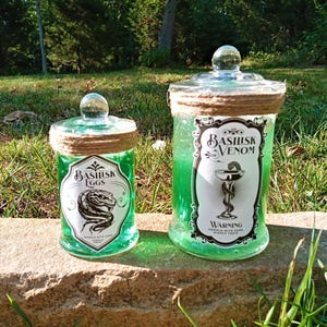 Puede incluir: Dos frascos de vidrio con líquido verde y etiquetas. El frasco más grande está etiquetado como "Veneno de Basilisk" y el frasco más pequeño está etiquetado como "Huevos de Basilisk". Ambos frascos tienen una cuerda marrón atada alrededor de la tapa.