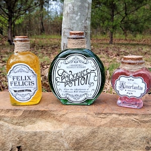May include: Three decorative potion bottles with cork stoppers. The bottles are filled with yellow, green, and red liquids. Each bottle has a label with text: "Felix Felicis", "Polyjuice Potion", and "Amortentia". The bottles are displayed on a stone surface.