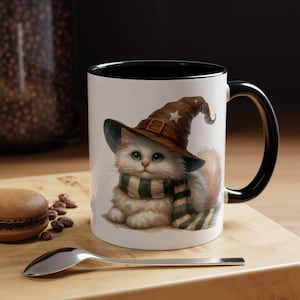 Puede incluir: Taza de cerámica blanca con interior y asa negros. La taza presenta una ilustración caprichosa de un gato blanco y esponjoso con un sombrero de mago marrón y una bufanda a rayas verdes y blancas. Una cuchara y un macaron están en primer plano.