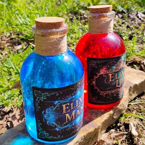 Könnte beinhalten: Zwei Glas-Trankflaschen mit Korken, eine gefüllt mit blauer Flüssigkeit und die andere mit roter. Jede Flasche hat ein Etikett mit den Worten "Elixir of Man" und "Elixir of Life". Die Flaschen sind mit Bindfaden umwickelt.