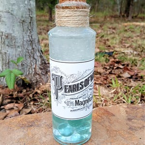 May include: A glass bottle with a cork stopper and a twine tied around the neck. The bottle is filled with a clear liquid and contains blue glass marbles. The label on the bottle reads "Pearls of Wisdom, Prepare and Lead, The Magnificence, In Search of Knowledge".