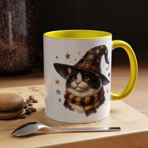 Puede incluir: Taza de cerámica blanca con interior y asa amarillos. La taza presenta una ilustración caprichosa de un gato blanco y negro con sombrero y bufanda de mago, con estrellas doradas. Una cuchara y un macaron están sobre una superficie de madera.