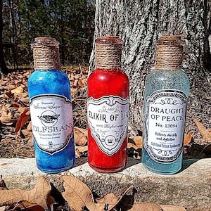 Könnte beinhalten: Drei Glasflaschen mit Korkstopfen und Etiketten. Die Flaschen sind mit blauer, roter und klarer Flüssigkeit gefüllt. Die Etiketten lauten "Wolfsbane", "Elixir of Life" und "Draught of Peace".