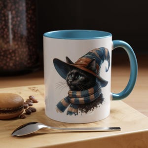 Puede incluir: Taza de cerámica blanca con interior y asa azules. La taza presenta un gato negro con un sombrero y bufanda de bruja con rayas azules y marrones. Una cuchara y un macaron están sobre una superficie de madera.