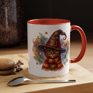 Puede incluir: Taza de cerámica blanca con interior y asa rojos. La taza presenta una ilustración caprichosa de un gato con sombrero y bufanda de mago, rodeado de estrellas y un fondo colorido. Una cuchara y un pastelito están en primer plano.