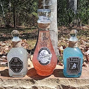 Könnte beinhalten: Drei dekorative Glasflaschen mit Korken und Etiketten. Die Flaschen sind mit schimmernden Flüssigkeiten in Rosa, Blau und Klar gefüllt. Auf den Etiketten steht "Cupid Crystals", "Tears of Mermaid" und "Pixie Dust".