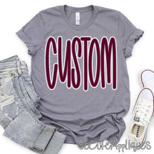 May include: A heather gray t-shirt with the word "CUSTOM" in a maroon color with a white outline. The shirt is paired with light wash denim jeans and white canvas sneakers.