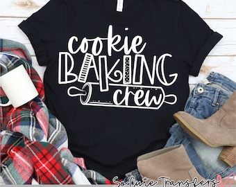 May include: A black t-shirt with white text that reads "cookie baking crew" and a rolling pin graphic. The shirt is on a wooden surface with a plaid blanket, a mug, and a pair of jeans.