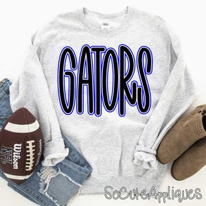 Può includere: Una felpa grigio chiaro con la scritta "GATORS" in grandi lettere nere con un contorno blu. Un pallone da football americano marrone e un paio di jeans blu sono accanto alla felpa. Il testo "SoCuteAppliques" è in basso a destra.