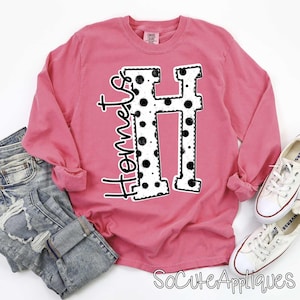 May include: A long-sleeved, pink shirt with the word "Hornets" in black script and a large, white letter "H" with black polka dots. The shirt is paired with blue jeans and white sneakers.