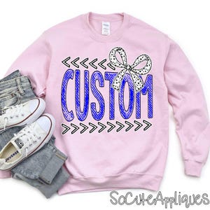 May include: A light pink sweatshirt with the word "CUSTOM" in blue, outlined in black, and a black and white polka dot bow. Black arrows frame the word. White sneakers and jeans are next to the sweatshirt.