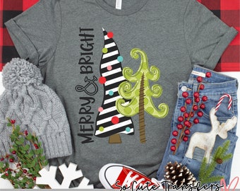 Op de afbeelding: Donkergrijs t-shirt met de tekst "MERRY & BRIGHT" en twee kerstboomontwerpen. Eén boom is zwart-wit gestreept met kleurrijke ornamenten, de andere is groen. Rode sneakers en een grijze muts zijn ook zichtbaar.