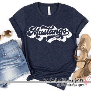 May include: Navy blue t-shirt with the word "Mustangs" in a white, distressed, retro font. The shirt is folded, with denim shorts, sunglasses, and sandals nearby. The shirt is from So Cute Transfers by SoCuteAppliques.