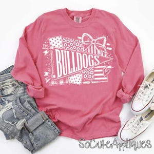 May include: A long-sleeve coral pink t-shirt featuring a white graphic print of a pennant flag with the text BULLDOGS, a large bow, and heart accents. The shirt is displayed alongside blue denim jeans and a pair of white canvas sneakers.