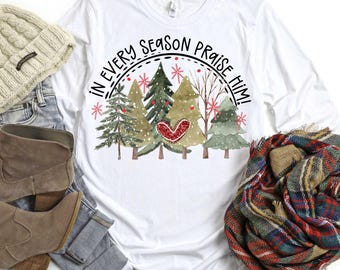 May include: White long-sleeve shirt with a graphic of Christmas trees and the text "IN EVERY SEASON PRAISE HIM!". The design includes a red heart and snowflakes. A beige beanie, jeans, boots, and a plaid scarf are also visible.