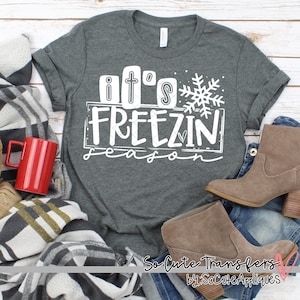 May include: A gray t-shirt with white text that reads "it's freezin season" with a snowflake graphic. The shirt is on a wooden surface with a blanket, a red mug, and brown boots.
