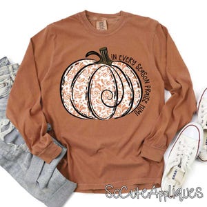 May include: A long-sleeved, rust-colored t-shirt with a white and orange pumpkin design. The text "In every season praise Him" is printed on the shirt.