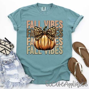 May include: A teal blue t-shirt with the text "Fall Vibes" repeated four times in a stacked design. A pumpkin with a leopard print bow is centered on the shirt.