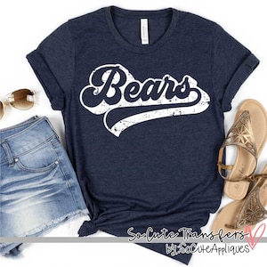 May include: A navy blue t-shirt with a white distressed vintage graphic that says "Bears" in a retro script font. The shirt is shown on a white background with a pair of sunglasses and a pair of sandals.