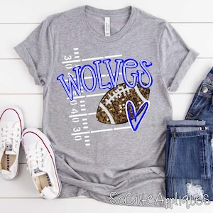 May include: A gray t-shirt with the word "WOLVES" in blue script, a football graphic, and a blue heart. The shirt also features a football field design. White sneakers and blue jeans are next to the shirt.