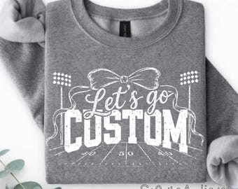 May include: A heather gray crewneck sweatshirt featuring a white graphic print. The design includes a large bow, stadium lights, a football field with yard lines, and the text Let's go CUSTOM in a distressed, bold, and script font combination.