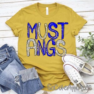 Può includere: T-shirt giallo senape con la scritta "MUSTANGS" in blu e lettere a pois neri. La maglietta è abbinata a jeans blu e scarpe da ginnastica bianche.